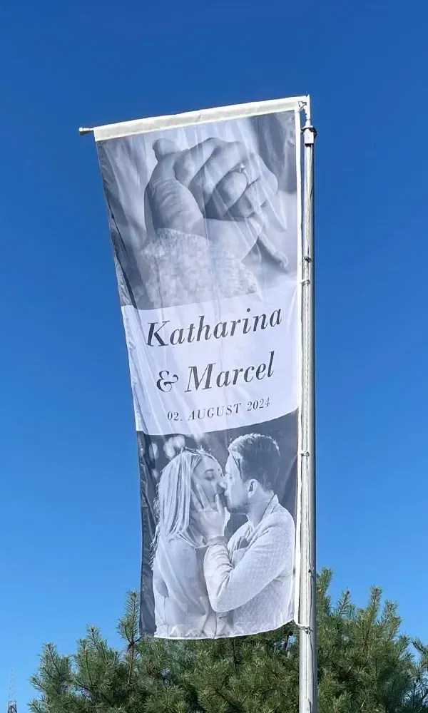 Hochzeitsflagge mit einem romantischen Bild von Katharina und Marcel, begleitet von ihrem Hochzeitsdatum „02. August 2024“, gehisst vor einem tiefblauen Himmel.