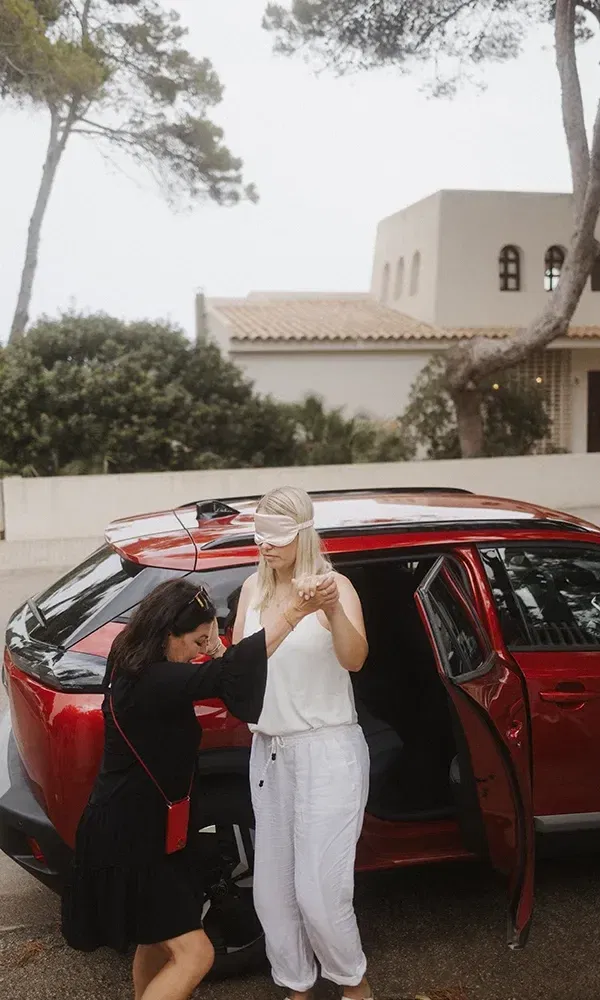 Eine blinde Überraschung: Eine Frau mit Augenbinde wird von einer Freundin vorsichtig aus einem Auto geleitet, mit einer Villa und Bäumen im Hintergrund.