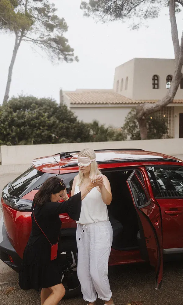 Eine blinde Überraschung: Eine Frau mit Augenbinde wird von einer Freundin vorsichtig aus einem Auto geleitet, mit einer Villa und Bäumen im Hintergrund.