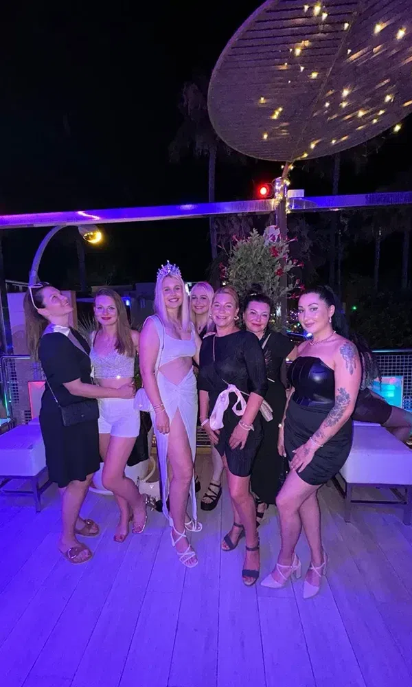 Sechs Frauen, darunter die Braut mit Krone und weißem Kleid, posieren zusammen auf einer abendlichen Feier unter funkelnden Lichtern.