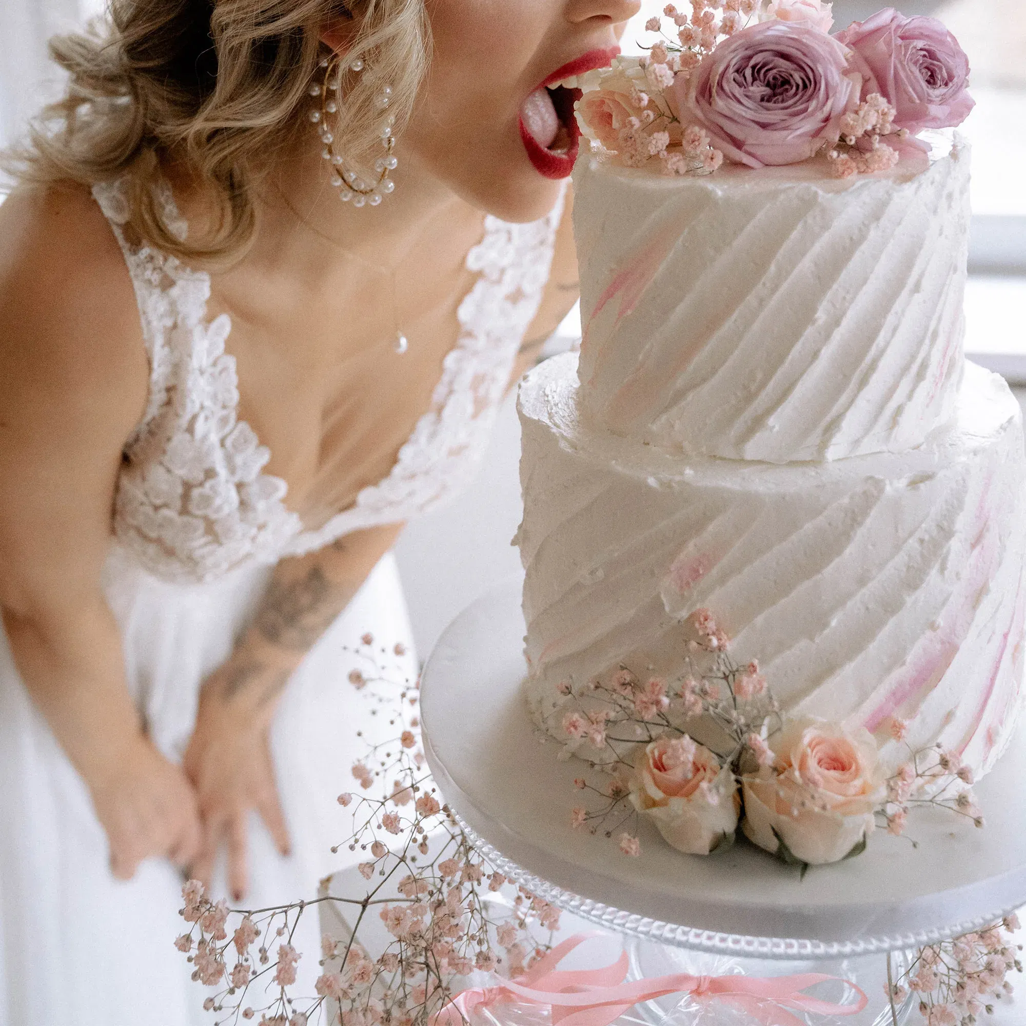 Eine Braut, die sich über eine zweistufige Hochzeitstorte beugt und spielerisch in die oberste Schicht beißt, umgeben von rosa und weißen Blumen.