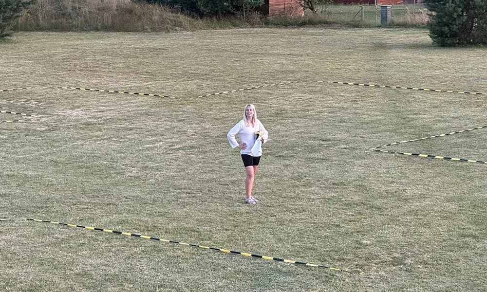 Person auf einem abgegrenzten Rasenbereich, umgeben von gelb-schwarzem Band, das den Standort für ein bevorstehendes Event markiert. Die Frau posiert fröhlich in sportlicher Kleidung.