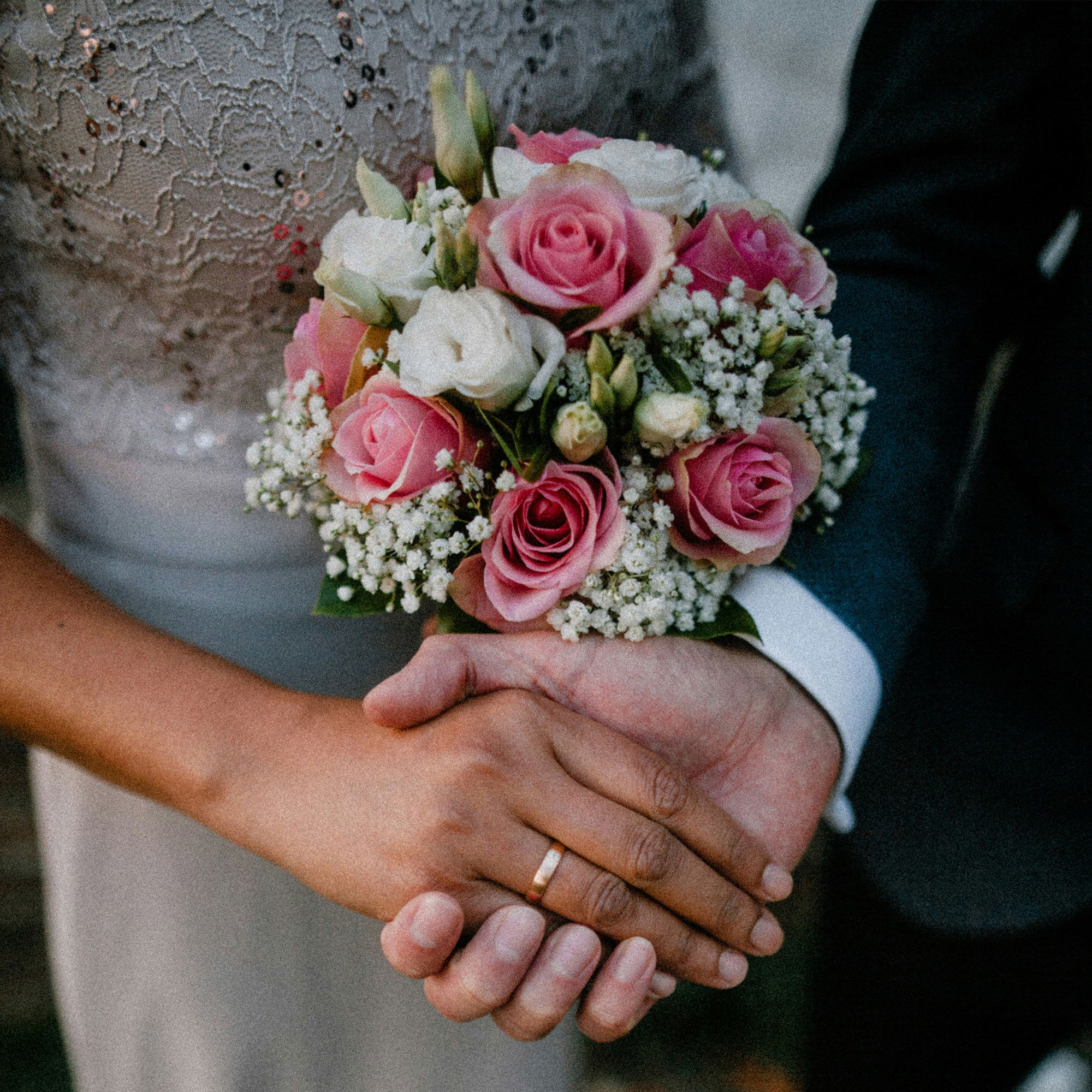 Bild von einem Brautpaar, das sich die Hände hält. Die Braut trägt ein graues Kleid mit Spitzenverzierungen und hält einen Blumenstrauß aus rosa und weißen Rosen sowie Schleierkraut. Der Bräutigam trägt einen dunklen Anzug. Beide tragen Eheringe.