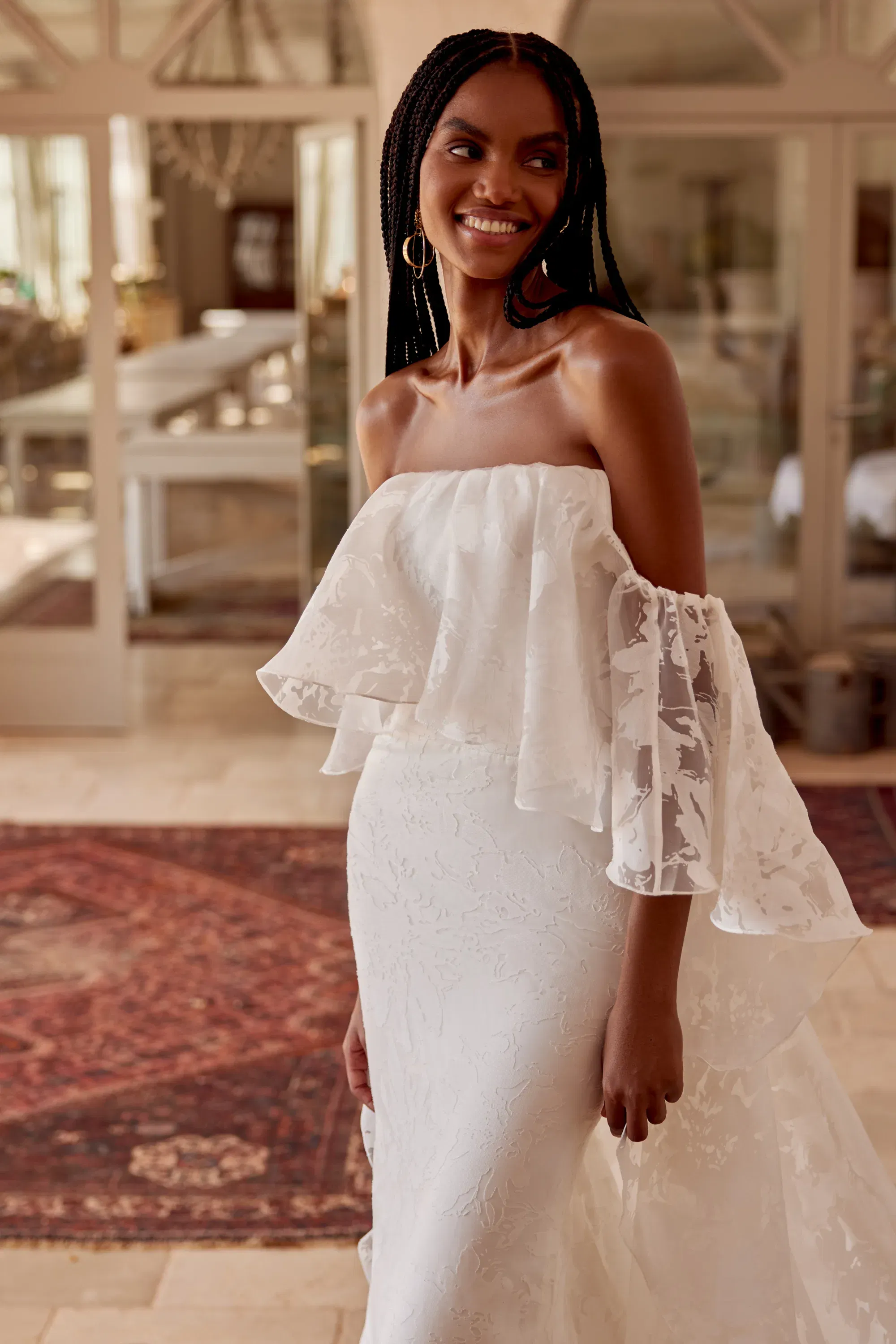 Nahaufnahme eines Brautkleides mit schulterfreiem Volant-Top und floraler Struktur, getragen von einem Model mit langen Braids.