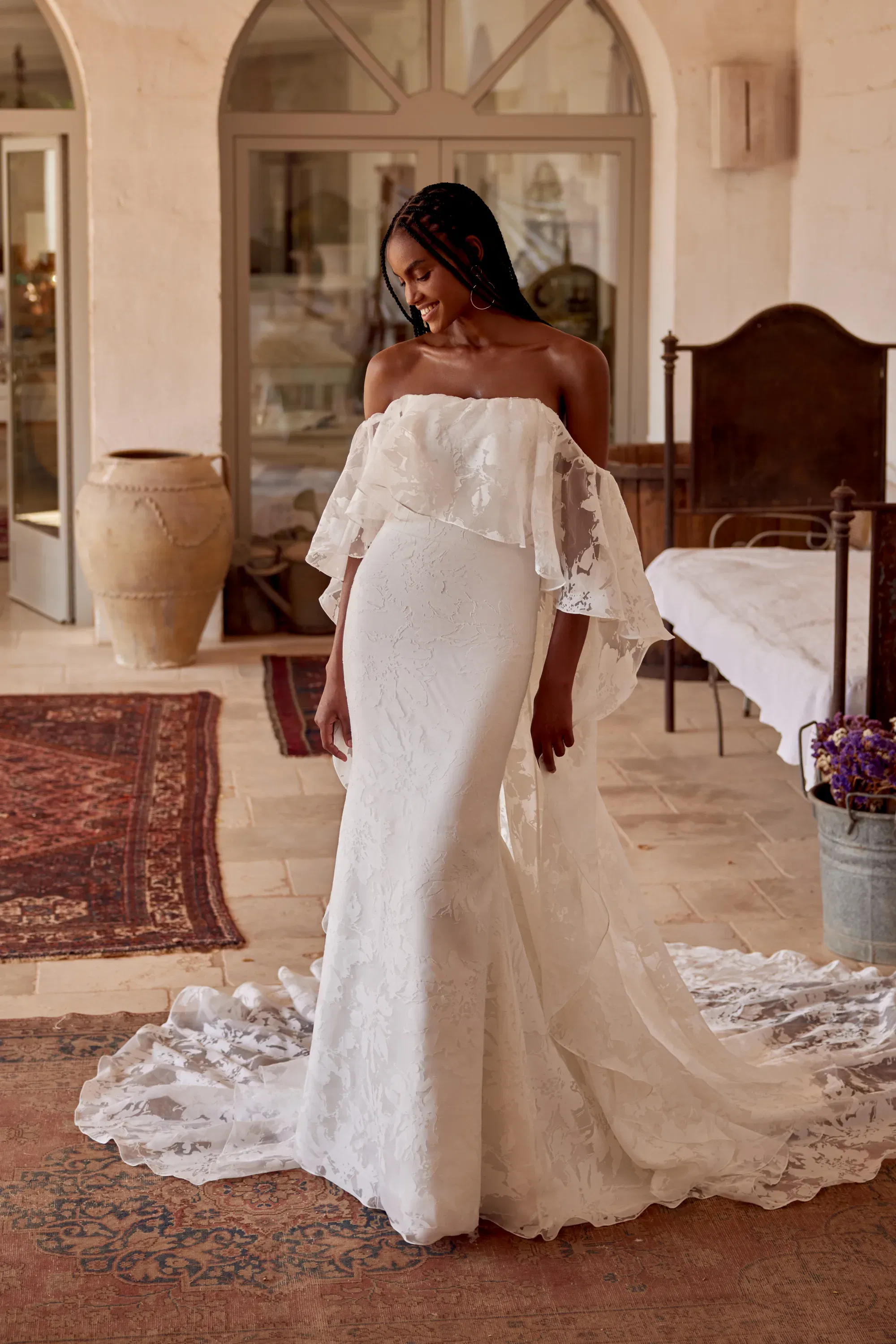 Ganzkörperaufnahme desselben schulterfreien Brautkleides mit langer Schleppe und floraler Textur, aufgenommen in mediterranem Innenhof.