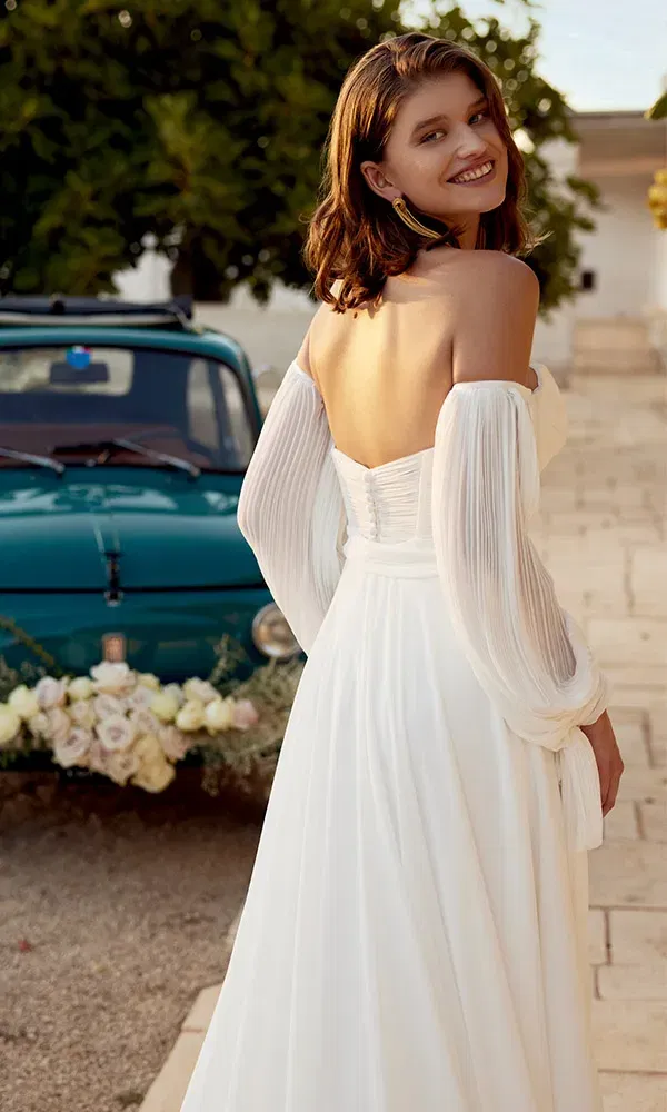 Rückansicht eines fließenden Boho-Brautkleides mit Schnürung und weiten Chiffonärmeln, Braut lächelt über die Schulter vor blauem Oldtimer