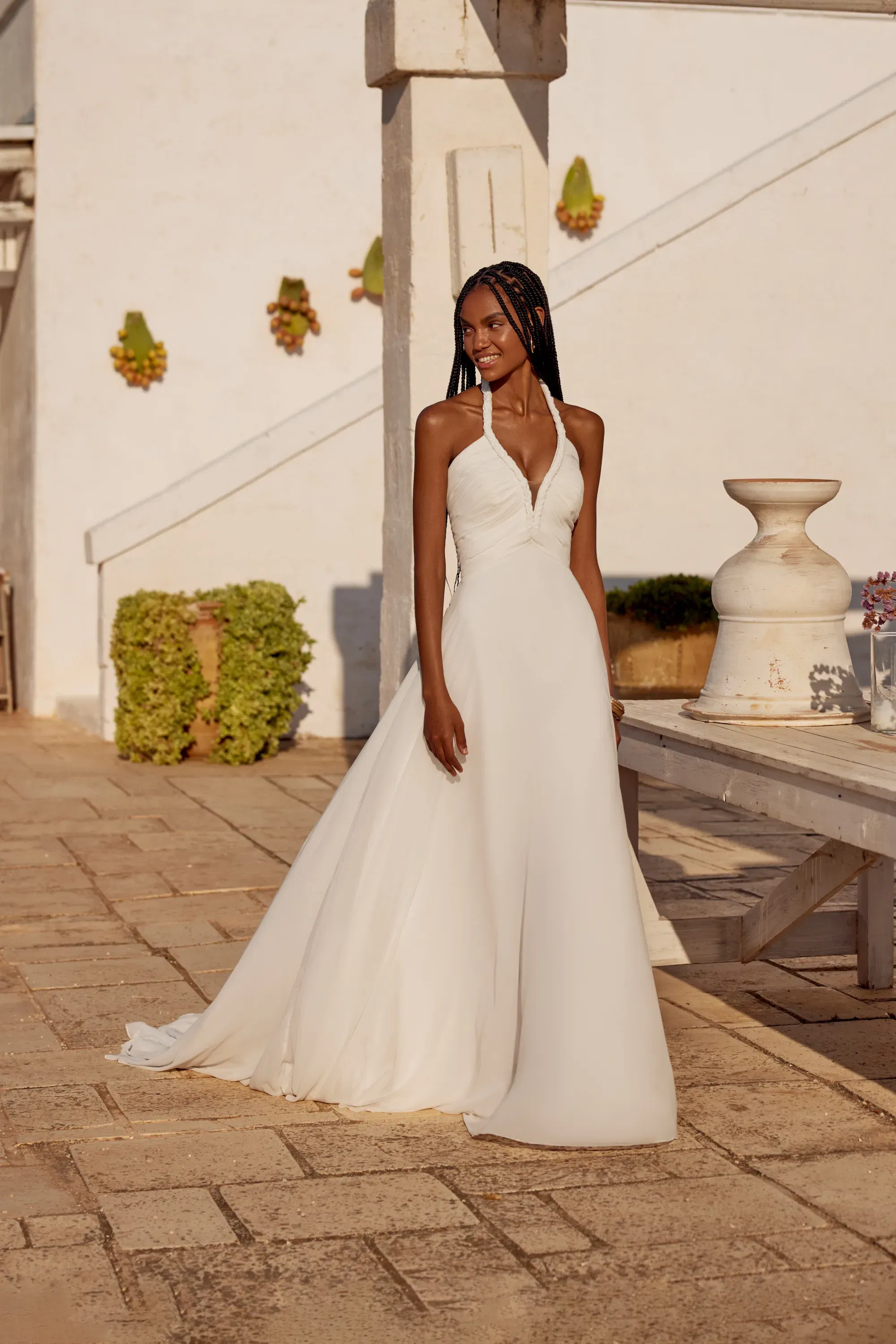 Ganzkörperaufnahme des gleichen Chiffon-Halterneck-Kleides mit ausgestelltem Rock, aufgenommen vor weißer mediterraner Kulisse.