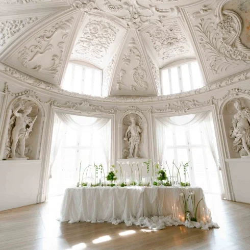 Elegant gedeckter Hochzeitstisch mit weißer Tischwäsche, zarten Blumenarrangements und Kerzen, inszeniert in einem prunkvollen Palais mit reich verzierten Stuckdecken, Skulpturen und großen Fenstern, die den Raum mit Tageslicht fluten.