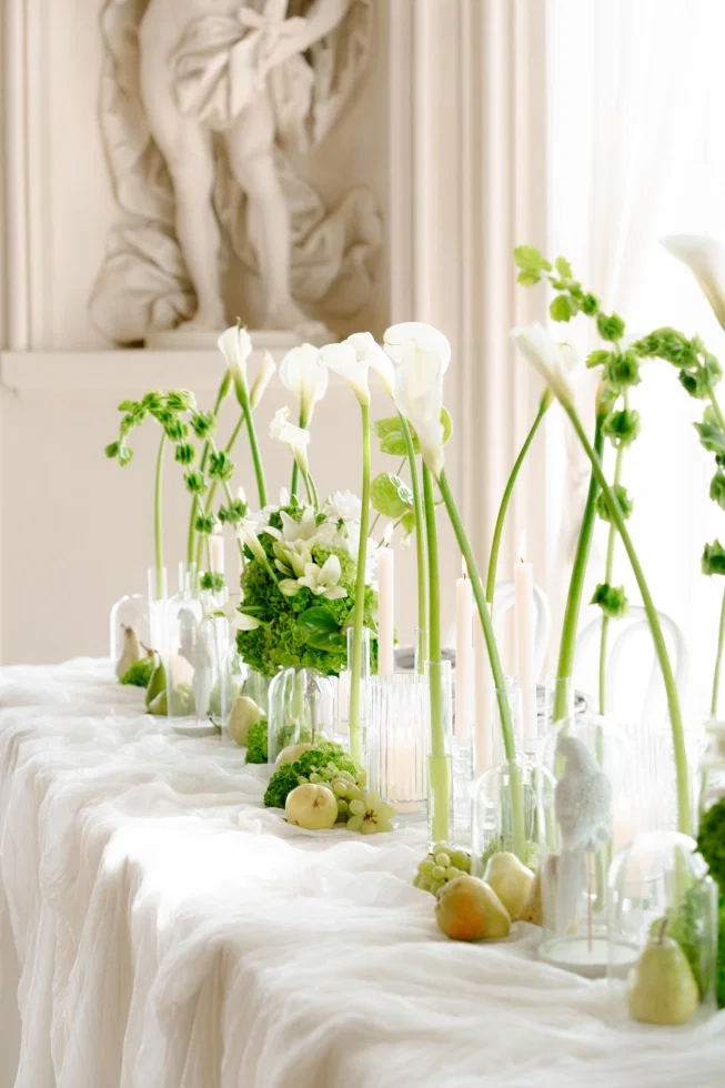 Elegante Hochzeitstafel mit weißen Calla-Lilien, grünen Blüten, Kerzen in Glasvasen und dekorativen Früchten auf fließendem Stoff.