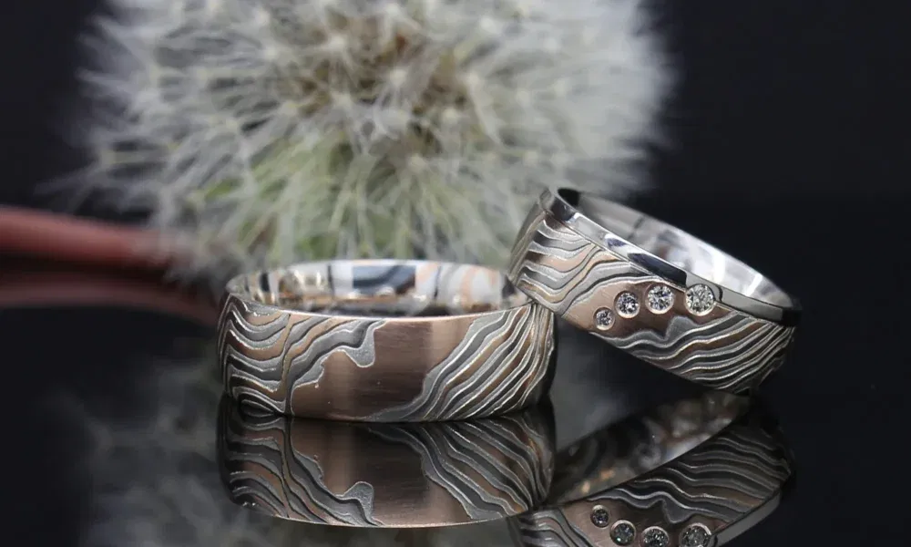 Zwei auffällige Eheringe aus Silber mit einem wellenförmigen, eingravierten Muster. Einer der Ringe ist mit mehreren kleinen Diamanten verziert. Im Hintergrund befindet sich eine Pusteblume, was dem Bild eine natürliche und elegante Atmosphäre verleiht.