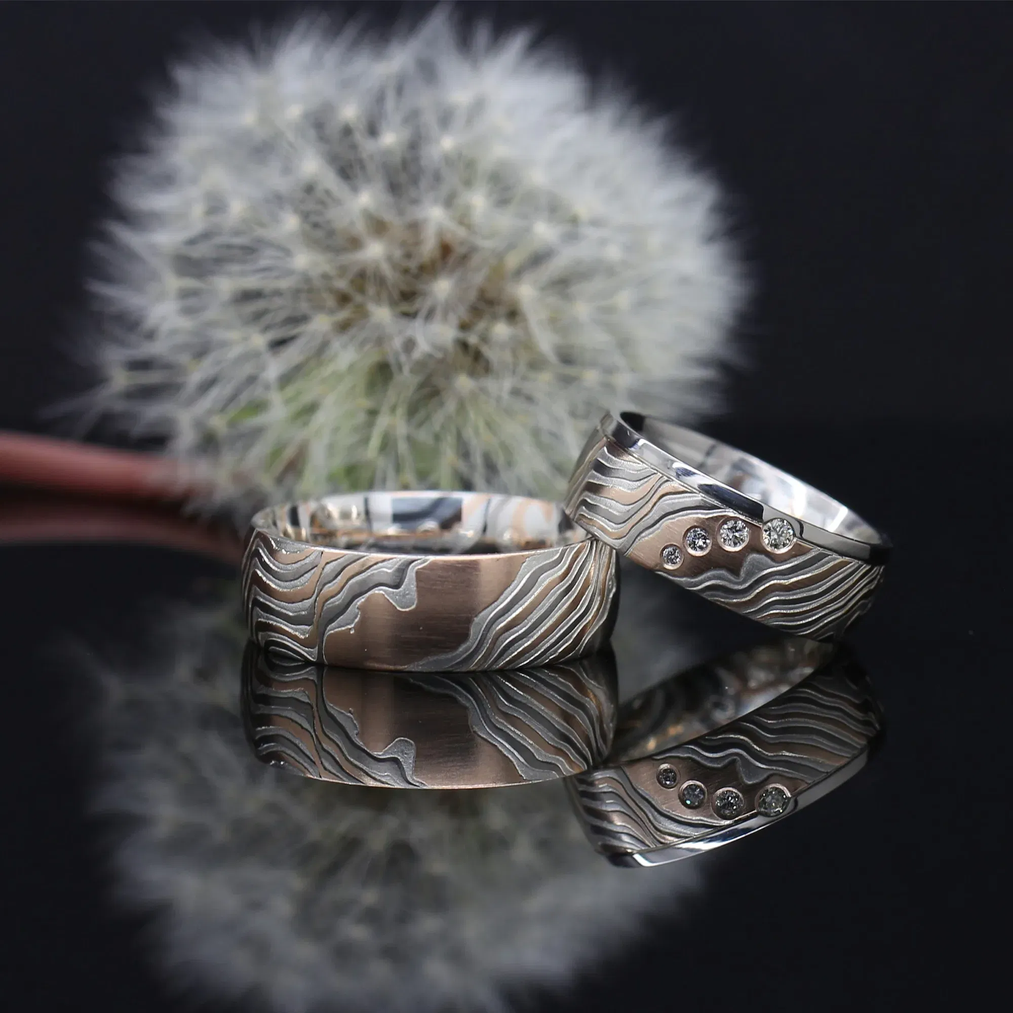 Zwei auffällige Eheringe aus Silber mit einem wellenförmigen, eingravierten Muster. Einer der Ringe ist mit mehreren kleinen Diamanten verziert. Im Hintergrund befindet sich eine Pusteblume, was dem Bild eine natürliche und elegante Atmosphäre verleiht.