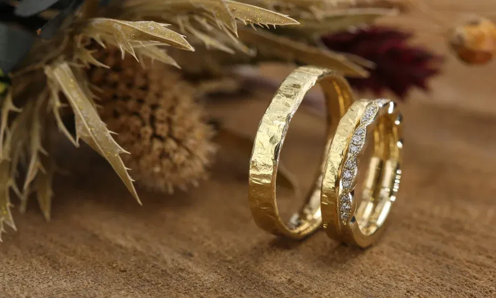 Handgefertigte, goldene Eheringe mit strukturierter Oberfläche, einer davon mit Diamanten besetzt, liegen auf einem Holztisch, umgeben von getrockneten Blumen.