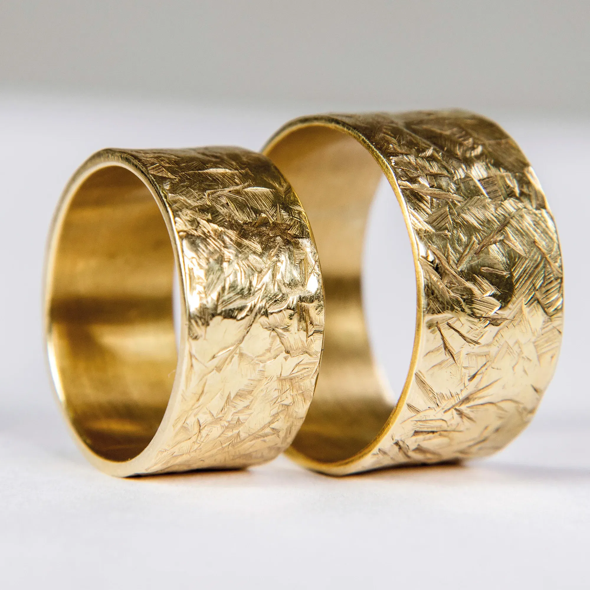 Zwei breite, goldene Eheringe mit handgehämmerter, strukturierter Oberfläche liegen auf weißem Hintergrund.