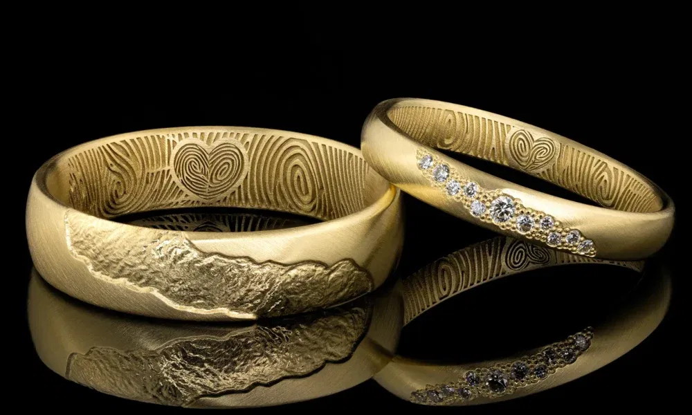 Goldene Trauringe mit handgravierten Details auf der Innenseite, einer mit einem Herz und Fingerabdruck, der andere mit kleinen Diamanten besetzt.