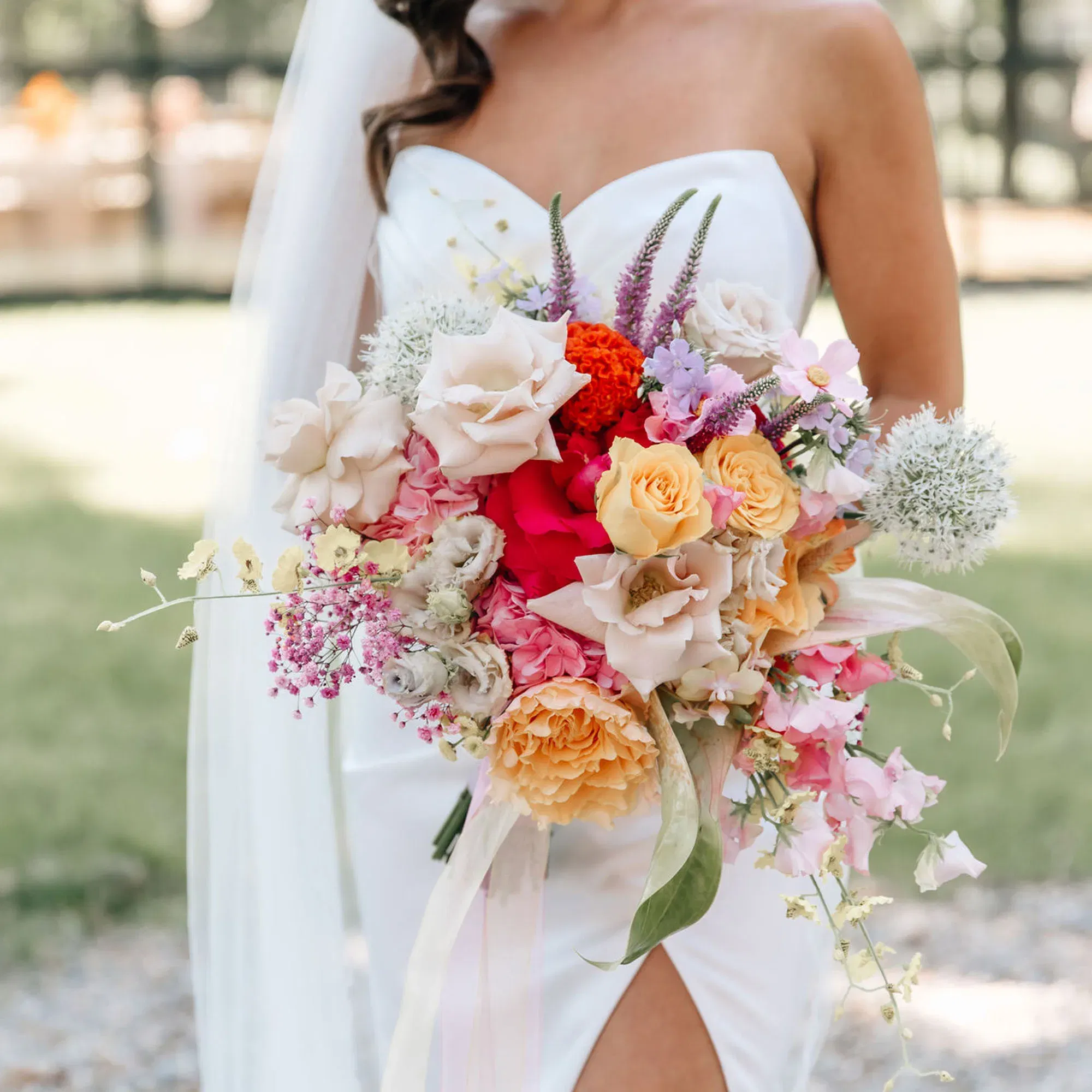 Die Braut trägt ein schulterfreies Kleid und hält einen detailreichen Blumenstrauß mit einer Mischung aus Rosen, pastellfarbenen Blüten und grünen Blättern. Im Hintergrund lässt sich eine unscharfe Gartenlandschaft erahnen.
