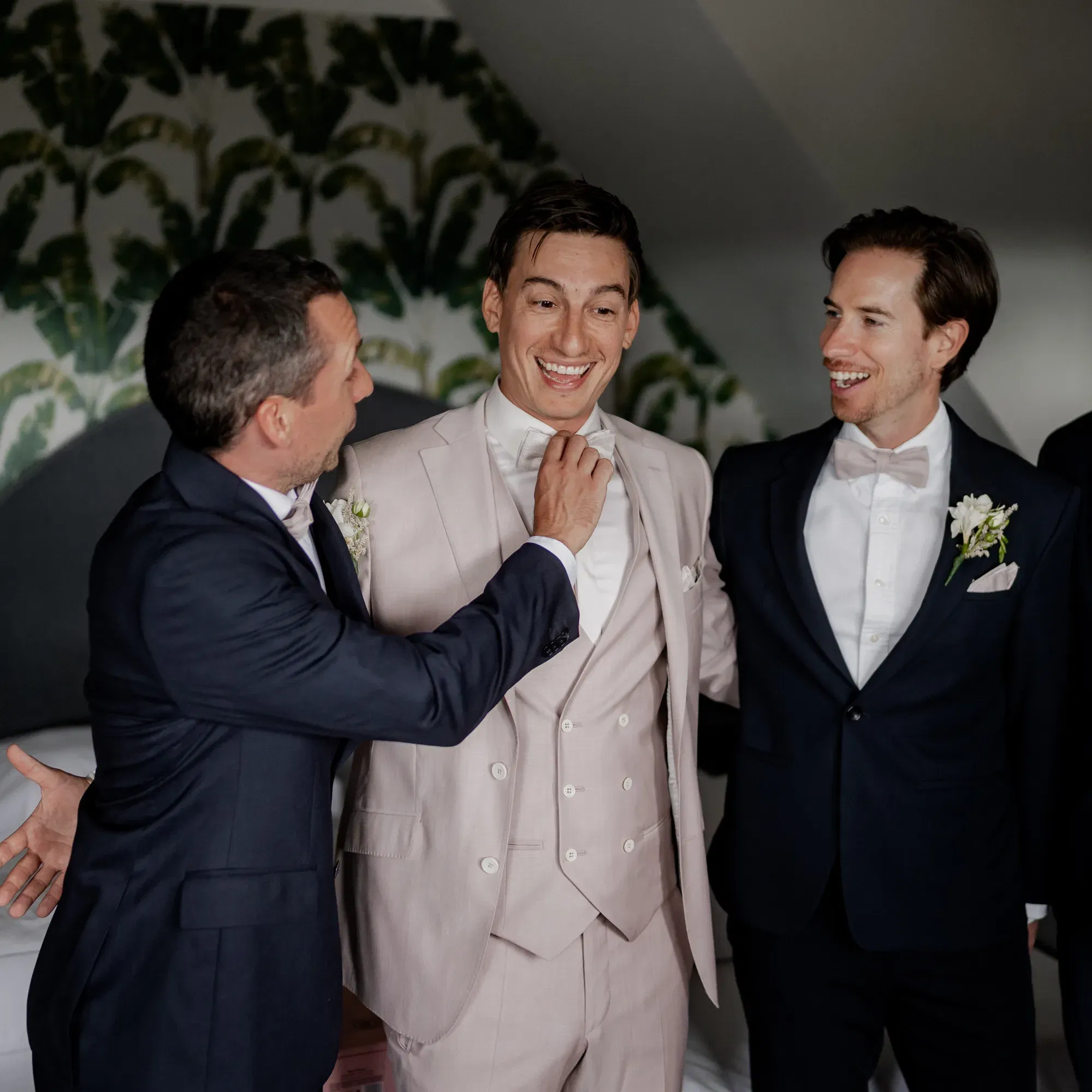 Der Bräutigam in einem cremefarbenen Anzug lacht, während zwei Groomsmen in dunklen Anzügen ihm bei den letzten Details helfen.