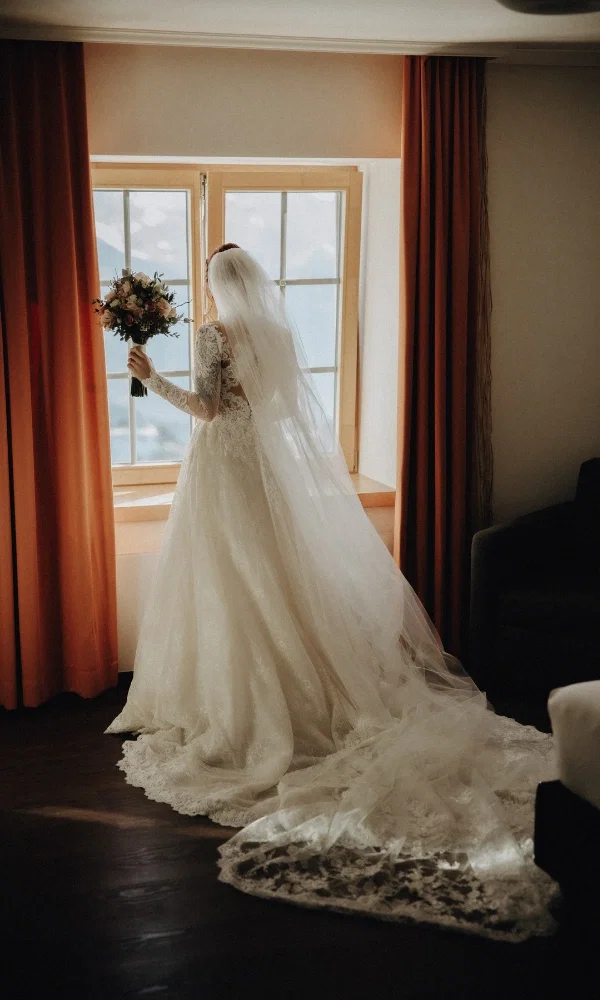 Die Braut schaut aus dem Fenster im weissen Hochzeitskleid und einem langen Schleier. Sie hält den Blumenstrauss in der Hand.