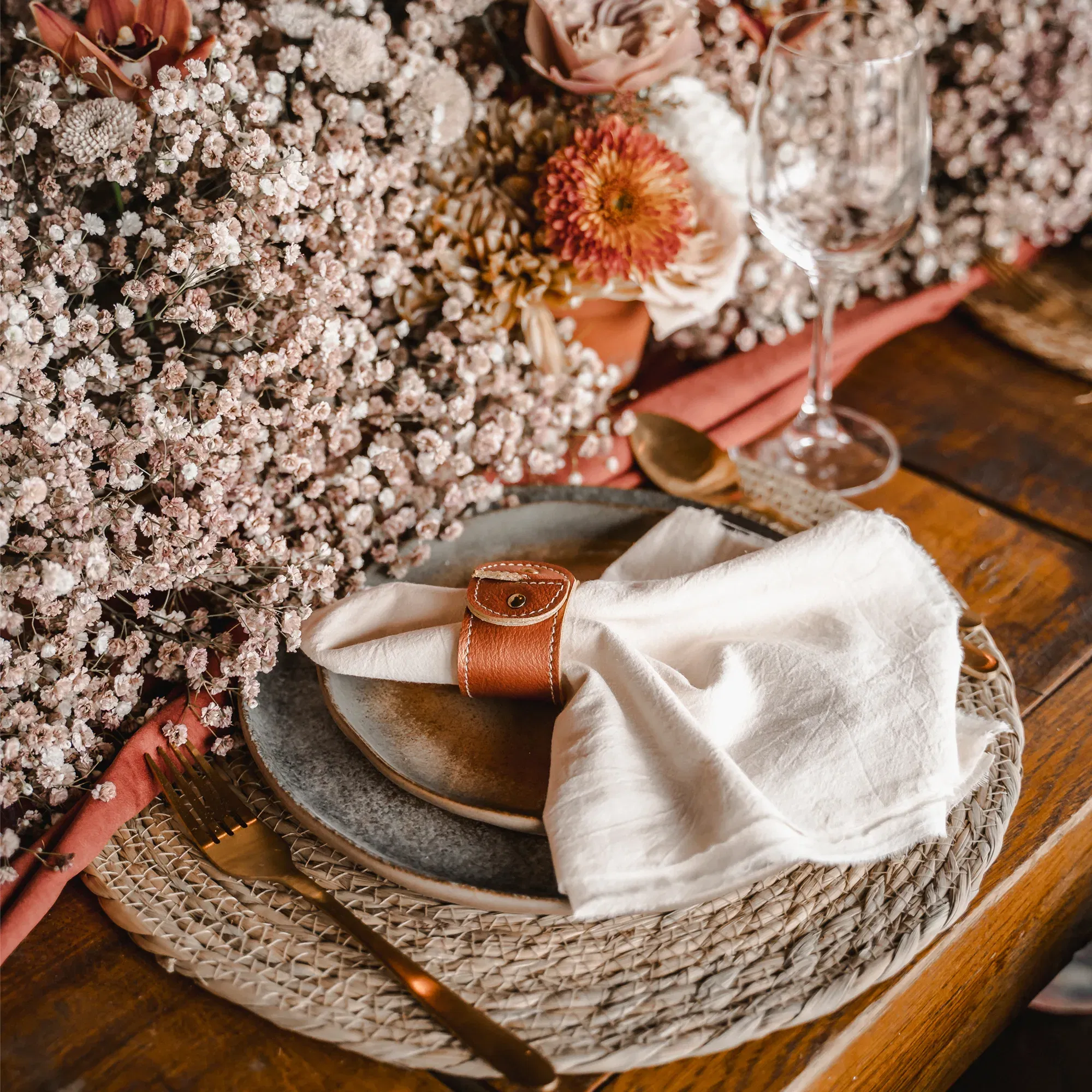 Natur-inspiriertes Hochzeitsdeko-Setting: Eine Serviette in einem Lederserviettenring auf einem rustikalen Teller, umgeben von üppigem Schleierkraut und Trockenblumen in zarten Farben.