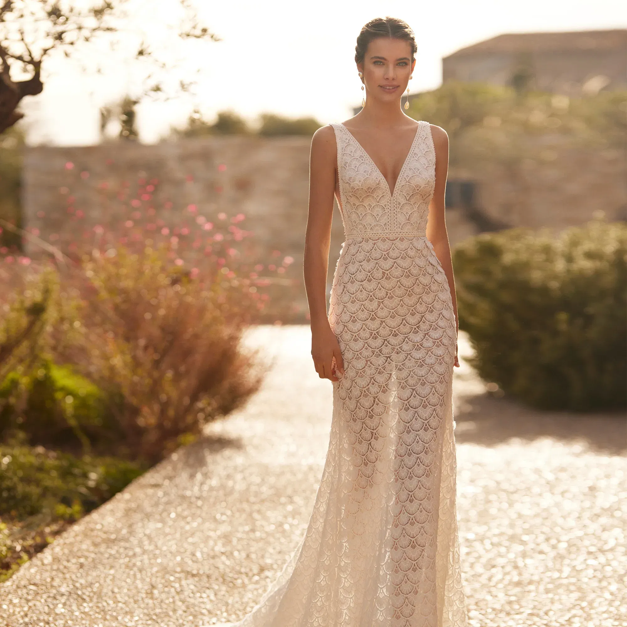 Braut im ärmellosen Spitzenkleid mit tiefem Ausschnitt, umgeben von einem sonnigen, blühenden Garten.