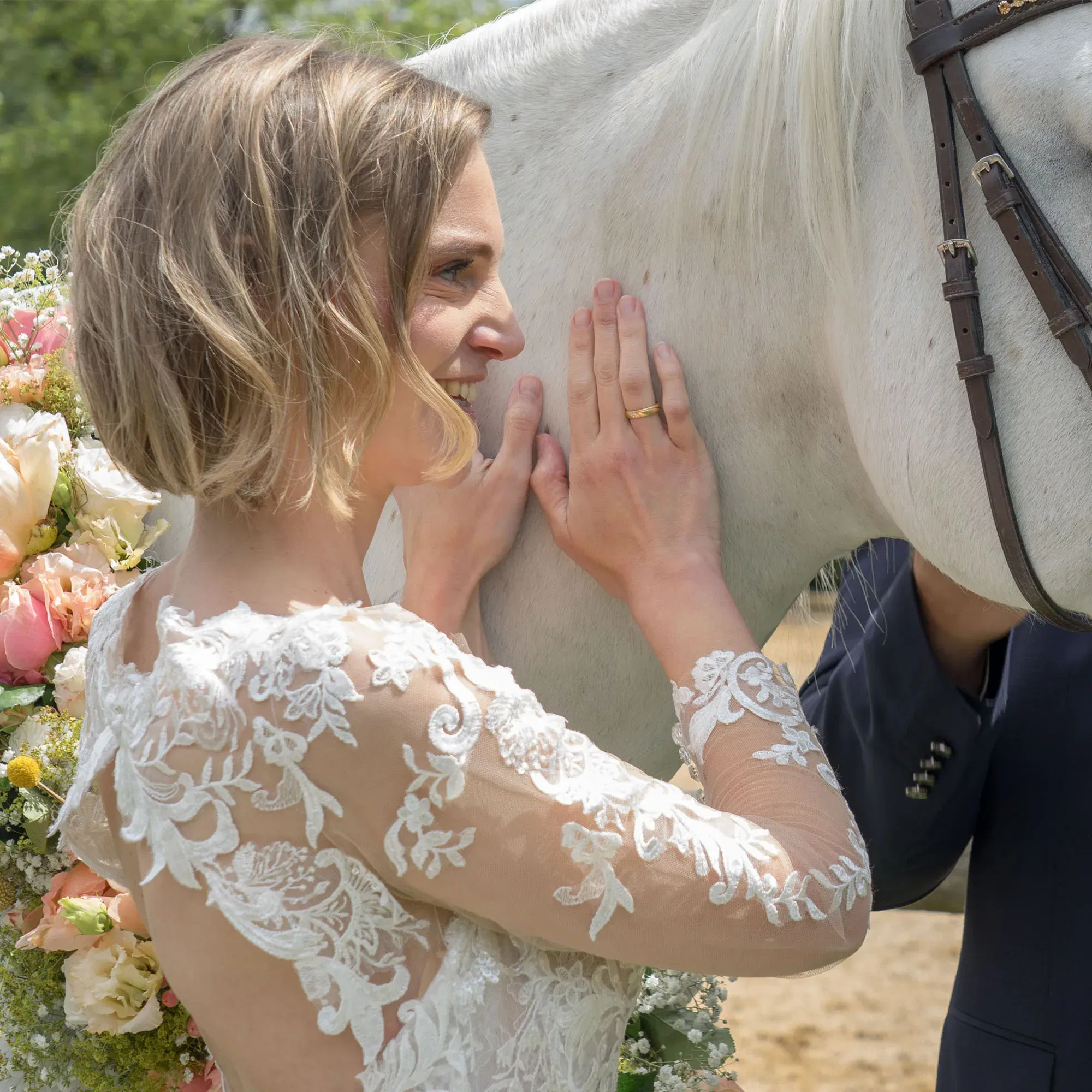 Eine Braut, die ein detailreiches, langärmeliges Spitzenkleid trägt, schmiegt sich lächelnd an den Hals eines weißen Pferdes und streichelt es liebevoll. Das Pferd ist mit einem Blumenkranz geschmückt, und die Szene findet auf einer Wiese statt.