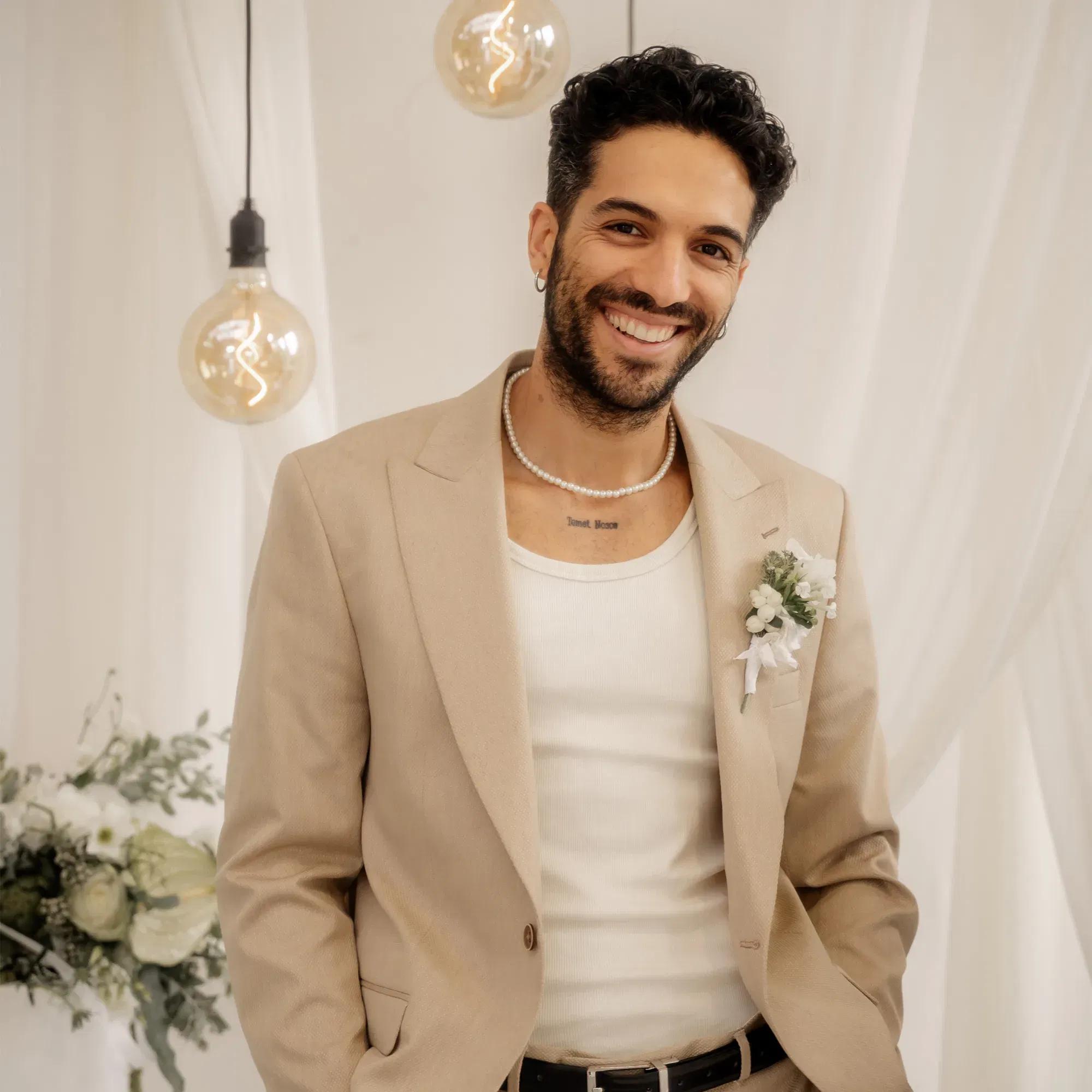 Lachender Mann im beigen Hochzeitsanzug, mit eleganter Boutonniere und Perlenschmuck, stehend vor einem dekorierten Hintergrund.