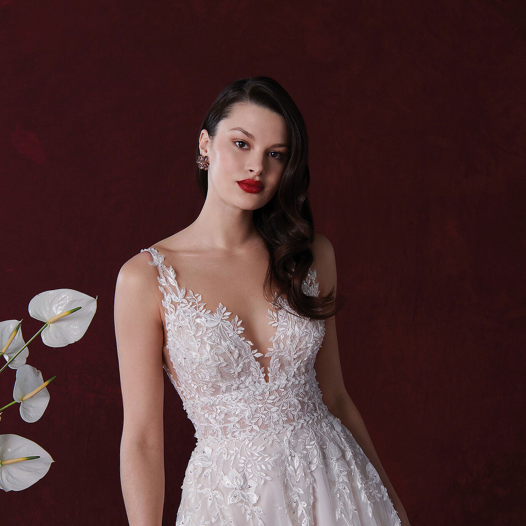 Elegante Braut in einem detailreichen Spitzenkleid von Justin Alexander, mit zartem floralen Design und tiefem Ausschnitt. Vor einem tiefroten Hintergrund posierend, trägt sie roten Lippenstift und hat ihre dunklen Haare in weichen Wellen gestylt.