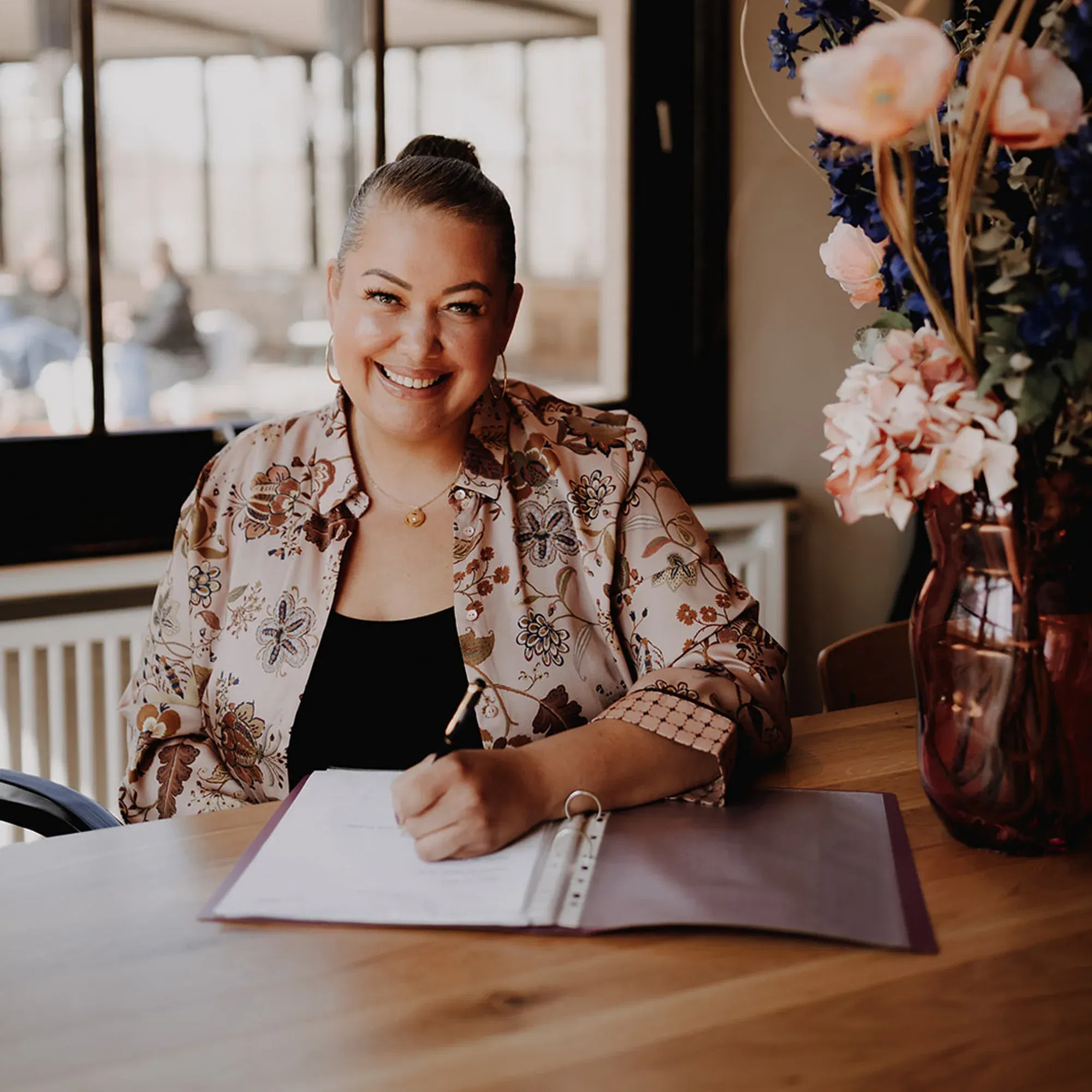 Eine lächelnde Frau mit einem ordentlichen Dutt sitzt an einem Tisch und arbeitet an einem Dokument. Sie trägt eine gemusterte Bluse, und neben ihr steht eine Vase mit bunten Blumenarrangements.
