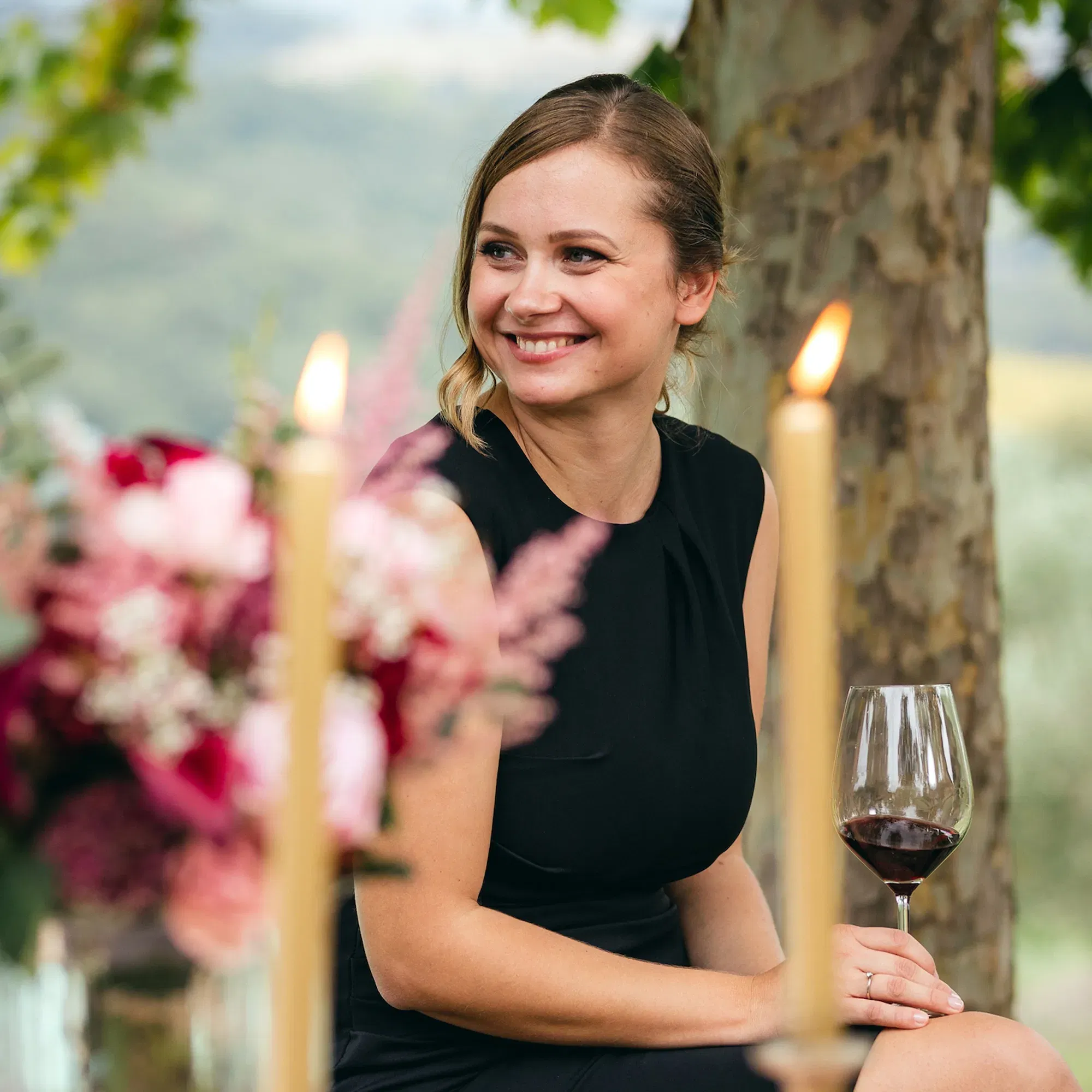 Eine lächelnde Frau in einem schwarzen Kleid sitzt draußen bei einem festlich gedeckten Tisch mit Kerzen und Blumenarrangements. Sie hält ein Glas Rotwein und schaut fröhlich zur Seite.