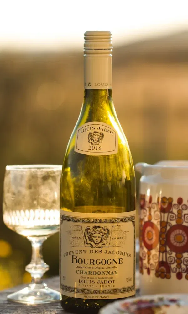 Stimmungsvolle Szene mit einer geöffneten Flasche Bourgogne-Wein, einem leeren Glas und einem bunt gemusterten Krug bei warmem Licht.