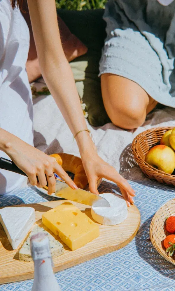 Nahaufnahme einer Hand, die Camembert auf einem Brett mit weiteren Käsesorten zerteilt, während andere Picknick-Elemente rundherum arrangiert sind.