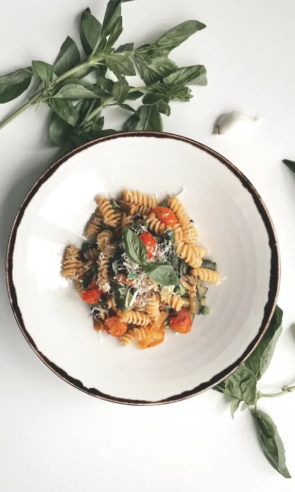 Mediterranes Pastagericht mit buntem Gemüse, serviert auf einem weißen Teller, garniert mit Basilikum und umgeben von Knoblauchzehen und Kräutern.