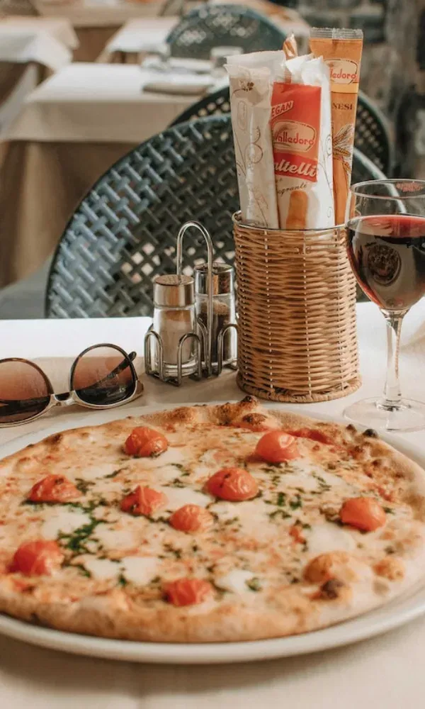 Einladend gedeckter Tisch mit einer Tomatenpizza, Gewürzspendern, Brotsticks, Rotwein und Sommeraccessoires – perfekte Urlaubsatmosphäre.