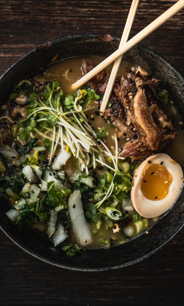 Herzhafte Ramen-Bowl mit aromatischer Brühe, Toppings wie Ei, Fleisch und knackigem Gemüse, angerichtet mit Stäbchen auf dunklem Holztisch.
