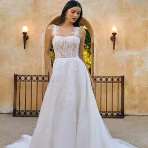 Braut in einem trägerlosen, figurbetonten Kleid mit floralen Spitzenverzierungen und transparenten Elementen, posiert in einem lichtdurchfluteten Raum.