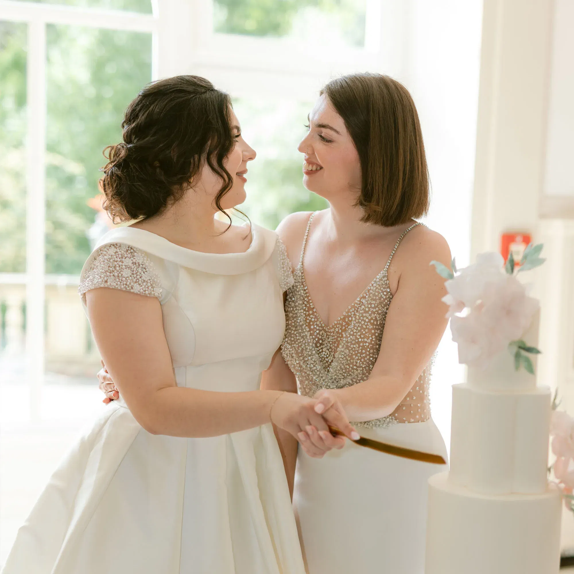Zwei glückliche Bräute schneiden gemeinsam ihre Hochzeitstorte an und strahlen dabei Freude aus. Die Braut links trägt ein schlichtes, weißes Kleid mit weitem Kragen, die Braut rechts ein funkelndes Kleid mit Perlenapplikationen. Durch die hohen Fenster fällt warmes Tageslicht, das die Szene erhellt.