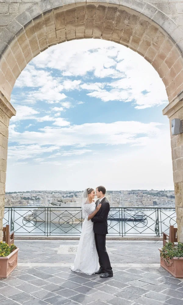 Ein Brautpaar steht innig umarmt unter einem historischen Bogen in Valletta, Malta, mit atemberaubendem Blick auf das Wasser und die Stadt im Hintergrund.