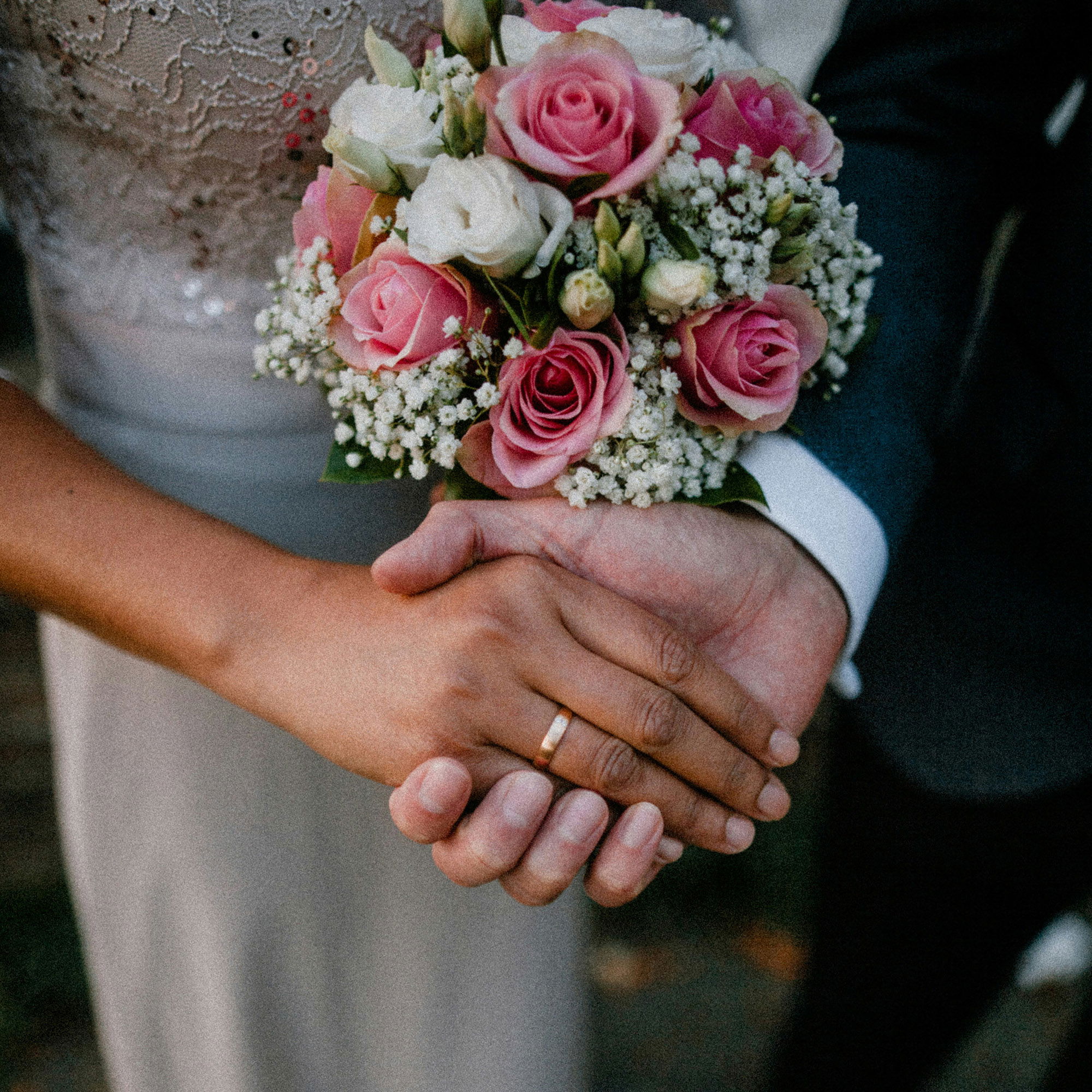 Ein Paar händchenhaltend, ihre Hand ist oben und man sieht ihren goldenen Ehering. In der anderen Hand hält sie einen Blumenstrauße mit weißen und rosanen Rosen.