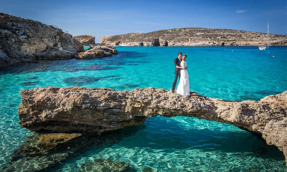 Braut und Bräutigam posieren auf einem natürlichen Felsen, der über das kristallklare, türkisblaue Wasser der Blue Lagoon auf Comino, Malta, ragt. Die Braut trägt ein elegantes Hochzeitskleid, der Bräutigam einen klassischen Anzug. Die umliegende felsige Küste und das ruhige Meer schaffen eine idyllische Hochzeitskulisse.