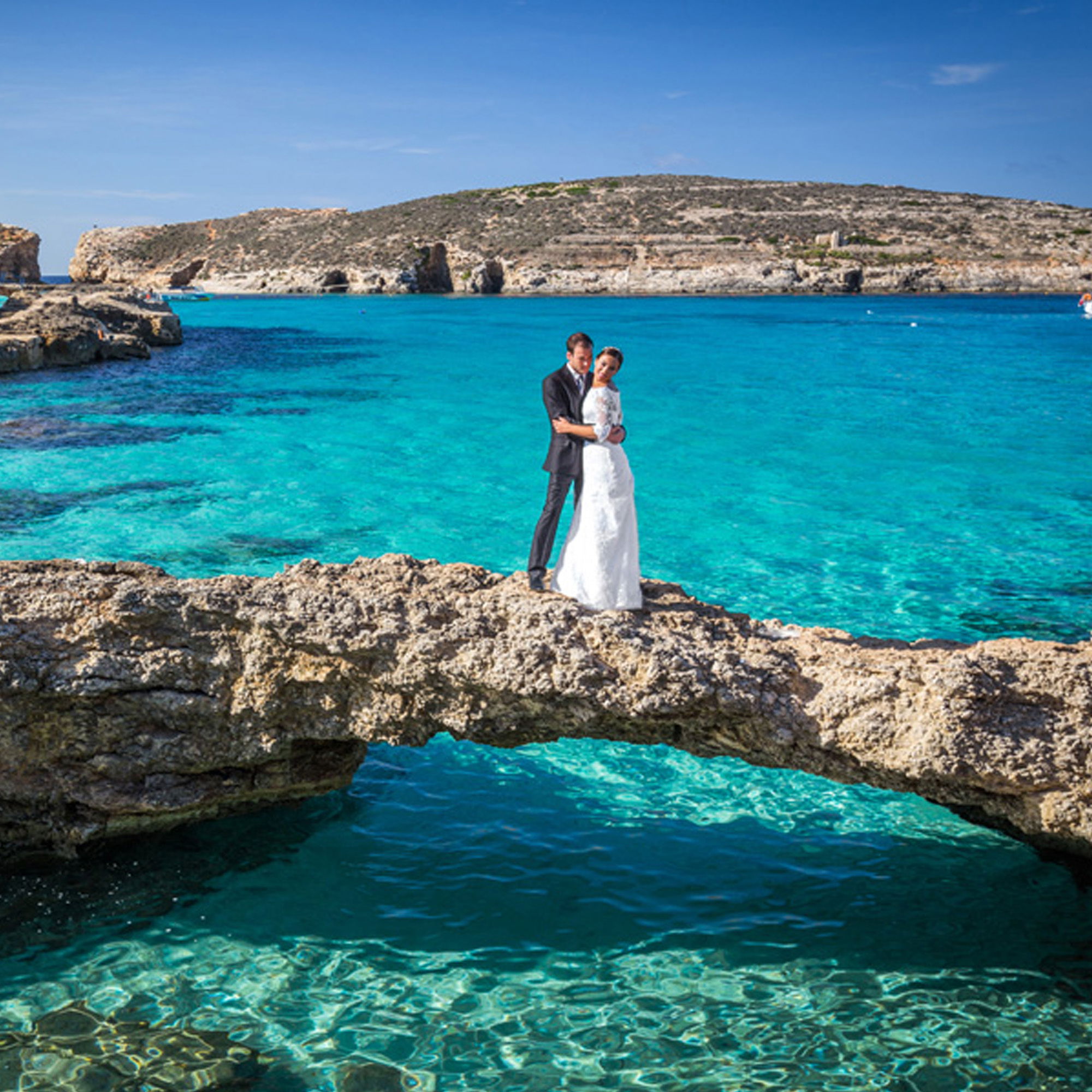 Braut und Bräutigam posieren auf einem Felsen mitten im kristallklaren, türkisblauen Wasser der Blue Lagoon, Comino, Malta. Die Braut in einem weißen Hochzeitskleid und der Bräutigam im dunklen Anzug stehen eng umschlungen auf einem natürlichen Steg, während die sonnenverwöhnte Felslandschaft im Hintergrund für ein romantisches Ambiente sorgt.