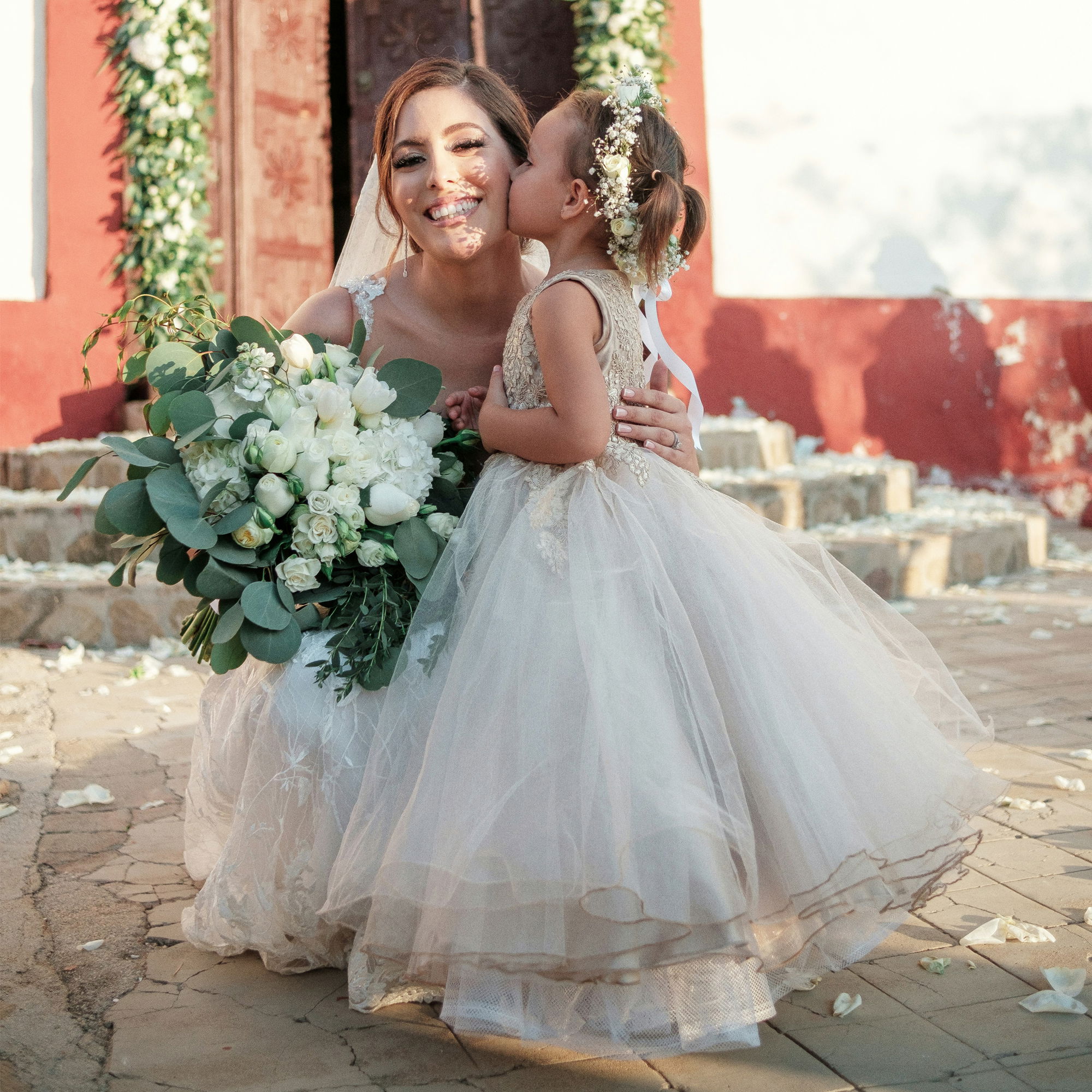 Junges Blumenmädchen in einem Tüllkleid küsst die Braut auf die Wange, beide in festlicher Kleidung und umgeben von Hochzeitsdekoration.