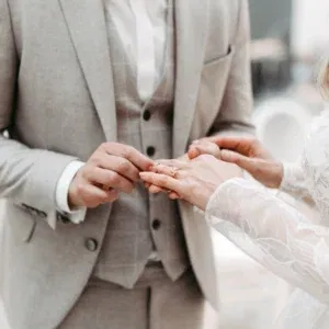 Nahaufnahme des Ringtausches: Der Bräutigam in einem grauen Anzug steckt der Braut den Ring an. Die Braut trägt ein weißes Kleid mit Spitzenärmeln, während der Moment in einer festlichen Umgebung stattfindet.