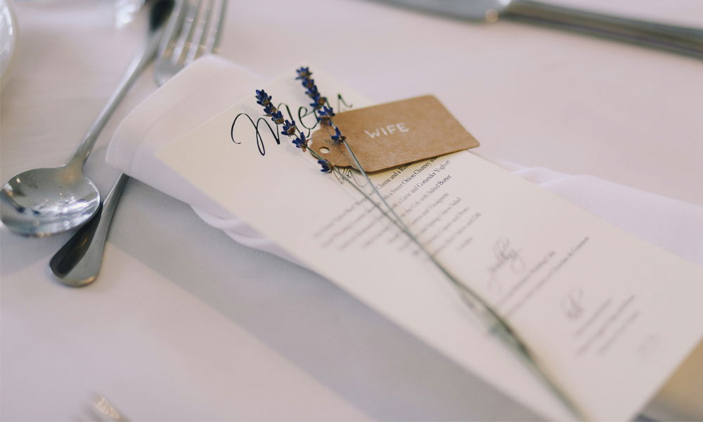 Hochzeitsgedeck mit einer eleganten Menükarte, die mit Lavendel und einem kleinen Namenskärtchen "WIFE" geschmückt ist. Die feine Tischdeko liegt auf einer weißen Stoffserviette, daneben glänzendes Besteck auf einer festlich gedeckten Tafel.