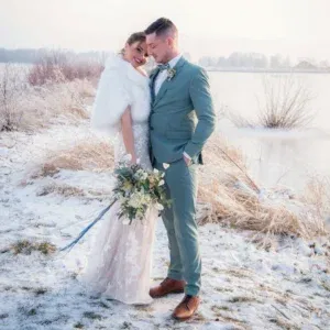 Brautpaar in einer verschneiten Landschaft an einem zugefrorenen See. Die Braut trägt ein elegantes Spitzenkleid und eine weiße Stola, während der Bräutigam einen stilvollen, hellblauen Anzug trägt. Die beiden strahlen Glück aus, während sie Arm in Arm stehen, und die Braut hält einen üppigen Blumenstrauß.