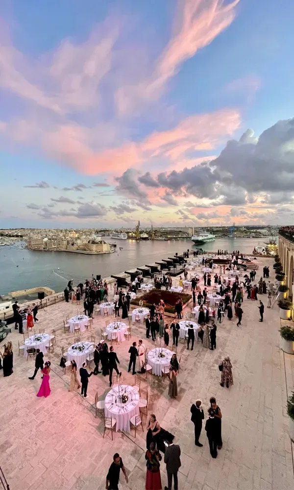 Hochzeitsfeier im Freien auf einer malerischen Terrasse in Valletta, Malta, mit festlich geschmückten Tischen, während der Himmel in sanften Rosa- und Blautönen über dem Hafen leuchtet.