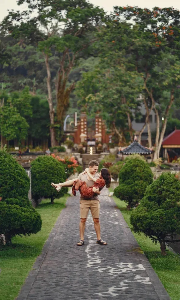 Verliebtes Paar vor fernöstlicher Tempelkulisse, er trägt sie durch einen Weg mit dekorativen Büschen und Bäumen.