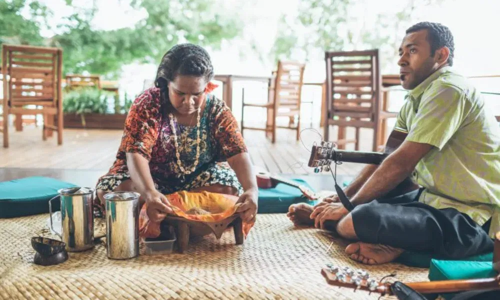 Zwei Personen sitzen auf Matten bei einer traditionellen Zeremonie, eine Frau bereitet ein Getränk in einem Tuch zu.