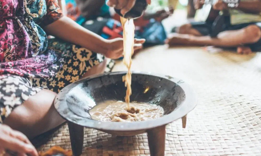 Nahaufnahme von Händen, die eine braune Flüssigkeit in eine Holzschale gießen – Teil einer traditionellen Getränkezubereitung.