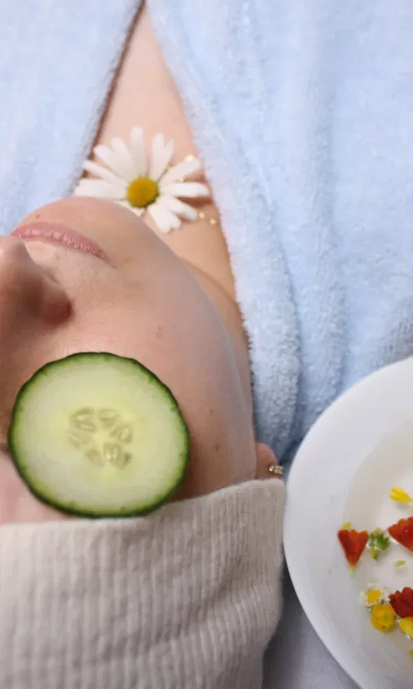 Entspannungsmoment im Spa: Frau in Bademantel mit Gurkenscheibe auf dem Auge, Blüte auf der Haut und Bowl mit Blüten und Snacks.