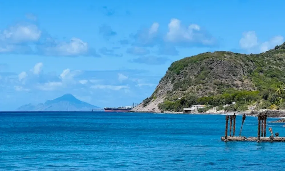 Inselpanorama mit sanften Wellen, einer kleinen Küstenansiedlung, einem alten Steg und einer Bergsilhouette am Horizont.