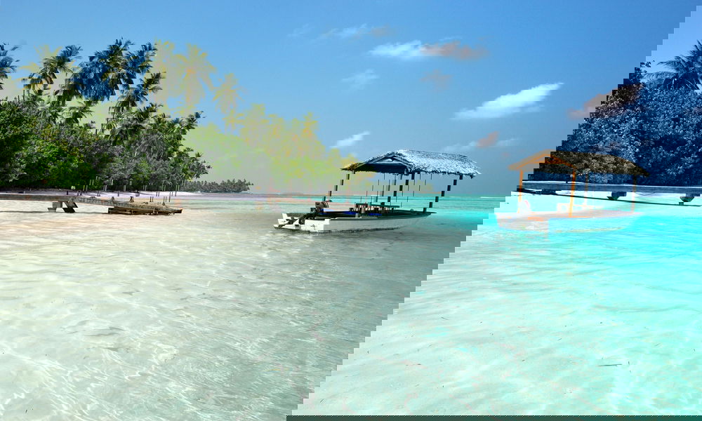 Ein kleines Holzboot mit Strohdach schwimmt im türkisfarbenen Wasser vor einem tropischen Strand mit Palmen und einem Holzsteg.